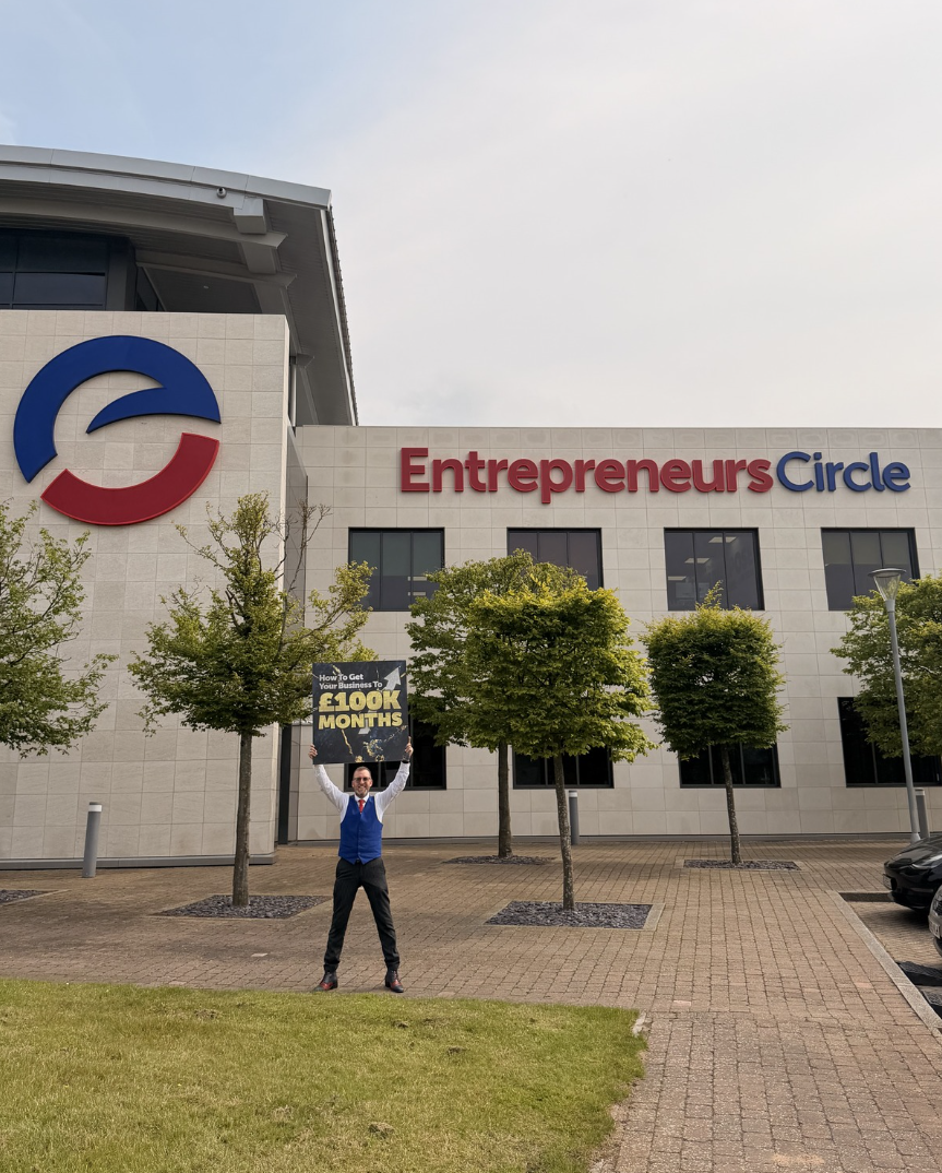 Nigel Botterill outside Entrepreneurs Circle HQ holding a How To Get Your Business To £100k Months sign